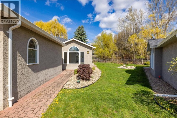 Side view of the townhouse showing a backyard with green grass, paved walkway, and trees. - 1020 Lanfranco Road Unit# 28, Kelowna