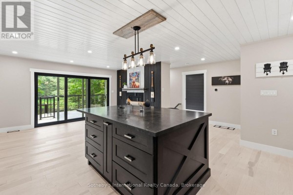 Open-concept living space with a dark island, fireplace, light wood floors, and sliding glass doors. - 1023 Strachan Point Road, Muskoka Lakes)