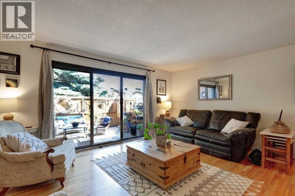 Living room with hardwood floors, furniture, and a large sliding glass door opening to a patio. - 103, 1305 Glenmore Trail Sw, Calgary