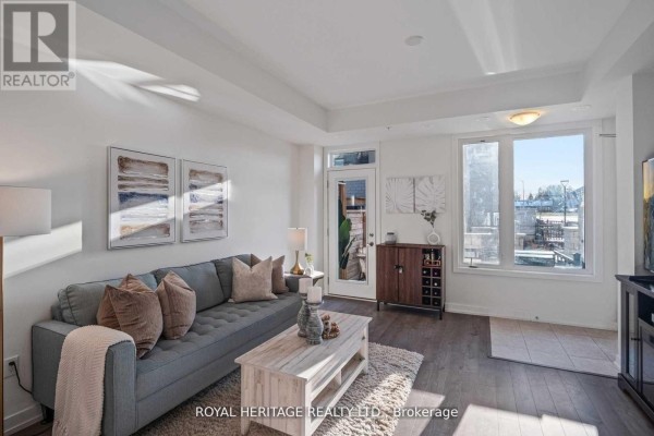 Living room with sofa, coffee table, view of windows, and door leading outside. - 103 - 1555 Kingston Road, Pickering