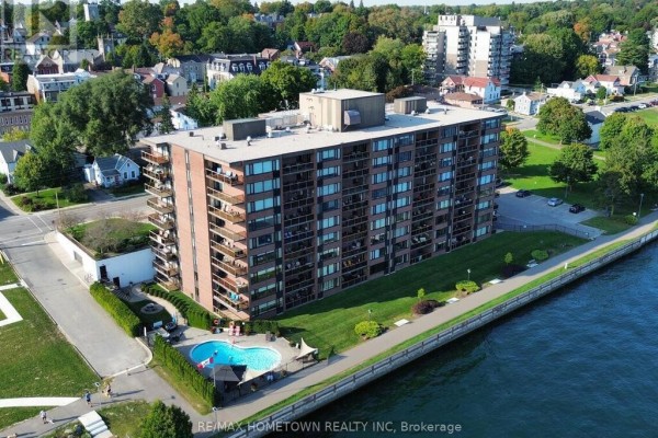 Aerial view of brick apartment building on waterfront with pool, green lawn, and surrounding trees. - 103 - 55 Water Street E, Brockville