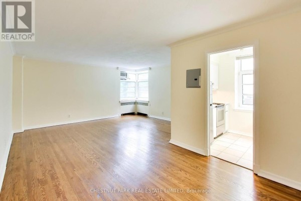 Empty living room with light beige walls, hardwood floors, and a corner window. - 103 - 717 Eglinton Avenue W, Toronto