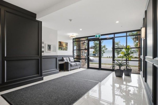 Building lobby with glass doors, seating area, plants, and dark wood paneling. - 103 2860 Trethewey Street, Abbotsford