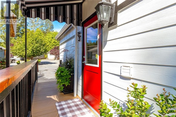 Close-up of a red front door with a black striped awning and wooden porch railing. - 103 3042 River Rd, Chemainus