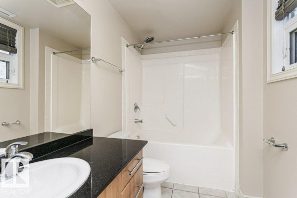 A bathroom with a white bathtub and shower, vanity with granite countertop, and toilet. - #103 9819 104 St Nw, Edmonton