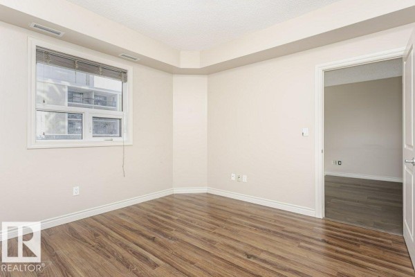 An empty bedroom with beige walls, wood-look laminate flooring, and a large window. - #103 9819 104 St Nw, Edmonton