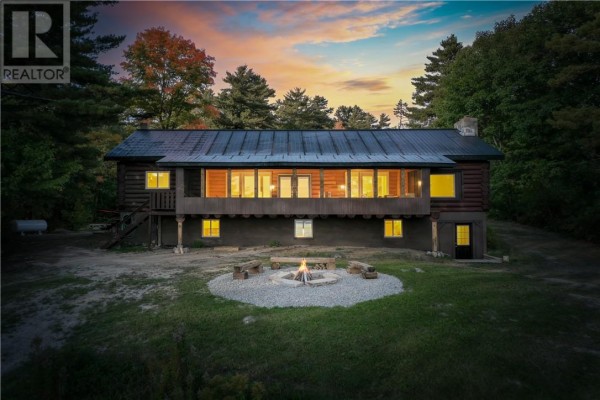 Log cabin at dusk with interior lights on and a lit fire pit. - 103a Northshore Road, Whitefish