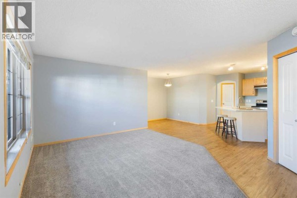 Open concept living room with grey carpet and a view into the kitchen with light wood cabinets. - 104, 23 Everridge Square Sw, Calgary