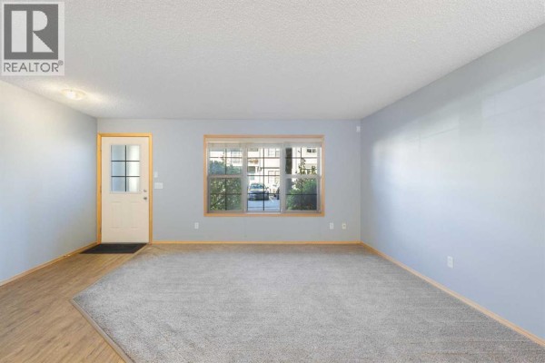 Living room with a white front door, grey carpet, and a window overlooking the parking lot. - 104, 23 Everridge Square Sw, Calgary