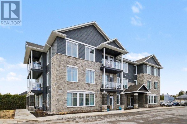 Exterior view of a modern condominium building with stone and dark siding during the day. - 104 - 5874 County Rd #19 Road, North Grenville