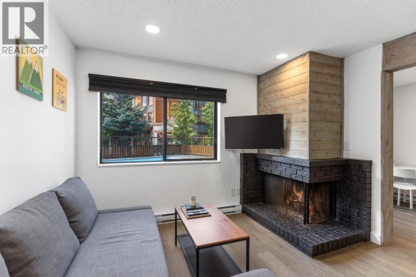 Living room with grey sofa, wood accent fireplace, and window view of apartment buildings and a pool. - 104 2129 Lake Placid Road, Whistler