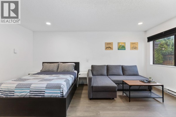 Studio bedroom with a queen bed, grey sectional sofa, coffee table, and window view of trees. - 104 2129 Lake Placid Road, Whistler