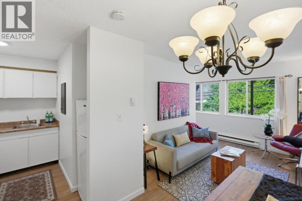 Compact kitchen with white cabinets and sink, adjacent to a living area with sofa and large windows. - 104 3465 Glen Drive, Vancouver