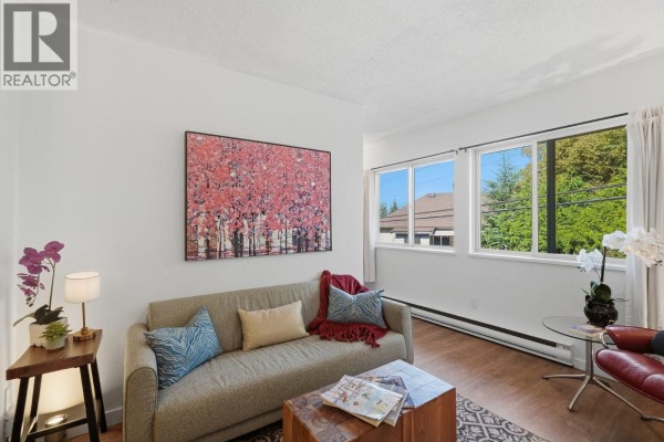 Living room sofa with colorful pillows and a large artwork, beside three windows with a tree view. - 104 3465 Glen Drive, Vancouver