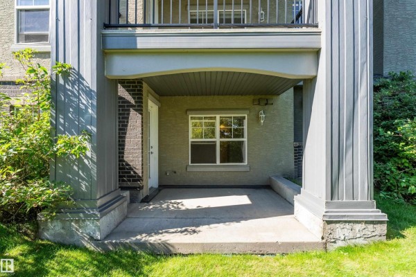 Covered concrete patio with pillars, a white door, a window, and surrounding greenery. - #104 630 Mcallister Lo Sw, Edmonton