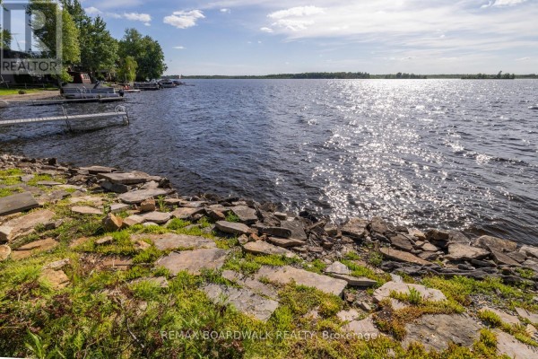 A rocky shoreline with green grass meets a wide, rippling lake under a bright sky. - 104 Avenue 4a, Beckwith
