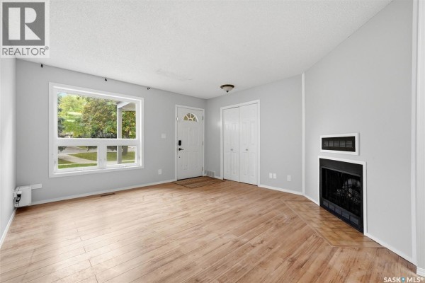 Living room with light wood laminate flooring, a large window overlooking green space, and a black gas fireplace. - 104 Cedar Meadow Drive, Regina