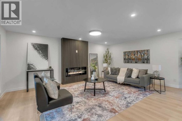 Living room featuring a modern fireplace, grey sofa, patterned rug, and floor-to-ceiling windows overlooking the backyard. - 104 Oakmoor Place Sw, Calgary
