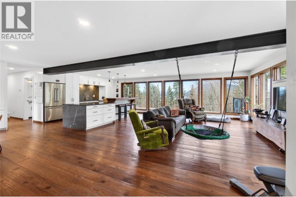 Living room with ample seating, large windows overlooking the pool, and dark built-in cabinets. - 1040 Old Auto Road Se, Salmon Arm