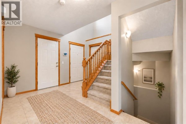 Interior foyer with beige tiled floors, carpeted stairs, wooden trim, and light grey walls. - 1041 Cranston Drive Se, Calgary