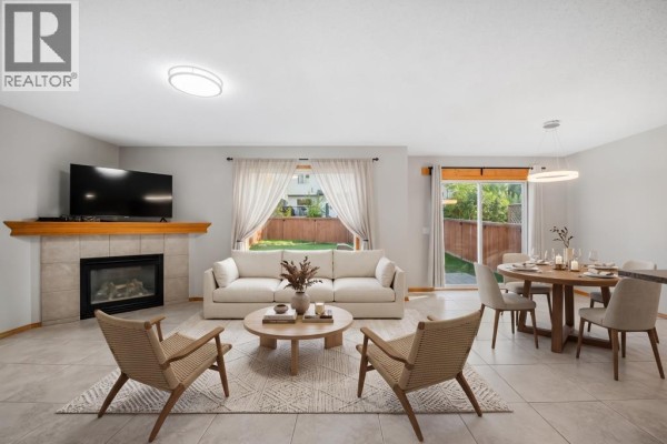 Living room with a fireplace, beige sofa, wooden coffee table, dining area, and backyard view through windows. - 1041 Cranston Drive Se, Calgary
