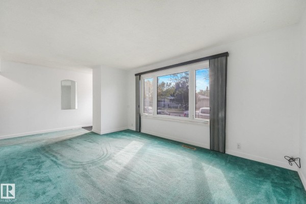 Living room with teal carpet, white walls, and a large window overlooking the street. - 10410 Lauder Av Nw, Edmonton