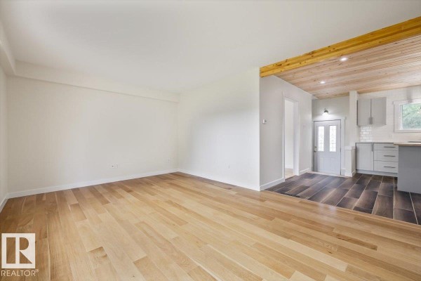 A wide view of the living room, showcasing light wood floors and a partial view of the kitchen with grey cabinets. - 10415 98 Av, Fort Saskatchewan