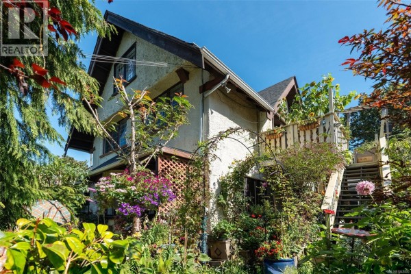 Side view of a stucco house with a wooden deck and vibrant flowering plants. - 1043/1045 Belmont Ave, Victoria
