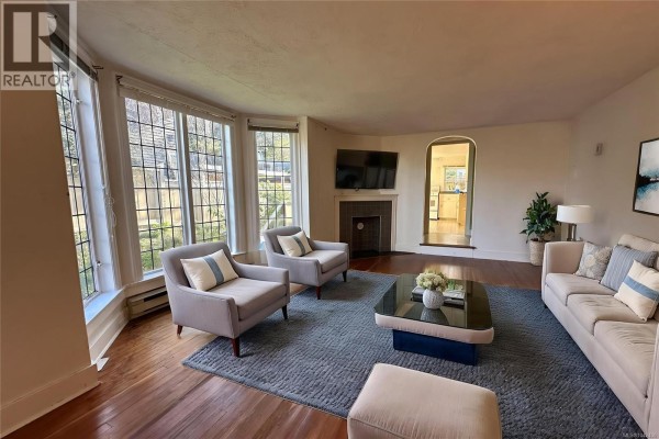 Living room with a large bay window, fireplace, sofa, chairs, and a coffee table. - 1043/1045 Belmont Ave, Victoria