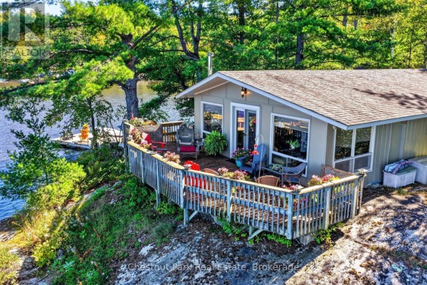 Cottage exterior with a wooden deck overlooking the water and surrounding trees. - 1048 Nora Lane, Gravenhurst