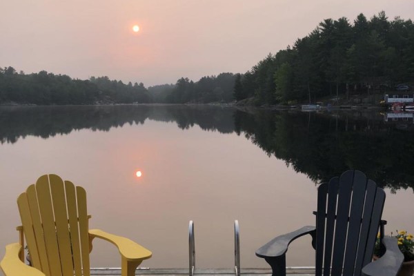 Two Adirondack chairs on a dock facing a hazy lake with two suns reflecting. - 1048 Nora Lane, Gravenhurst