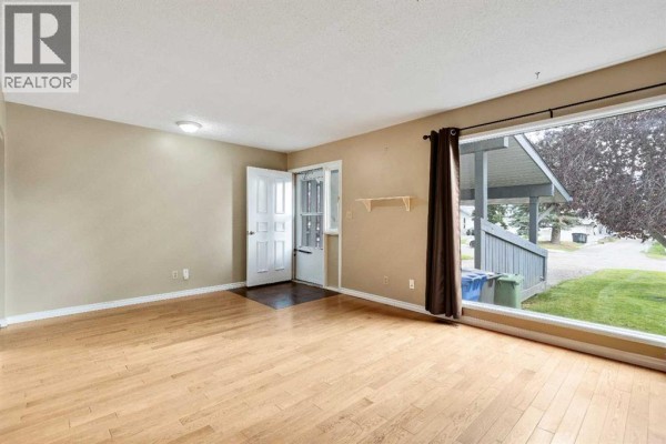 Living room with light wood floors, an open front door, and a large window showing the exterior. - 105, 107 Elm Place, Okotoks