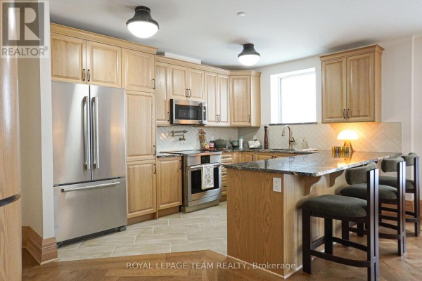Modern kitchen with light wood cabinets, stainless steel appliances, granite countertops, and a breakfast bar with stools. - 105 - 1050 Canadian Shield Avenue, Ottawa