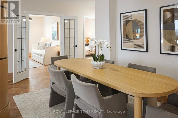 Dining room with a light wood table, grey upholstered chairs, decorative artwork, and glass-paneled doors to bedrooms. - 105 - 1050 Canadian Shield Avenue, Ottawa