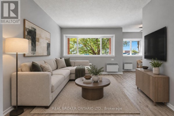 Living room with a sectional sofa, coffee table, and large windows overlooking trees. - 105 - 2044 Arrowsmith Drive, Ottawa
