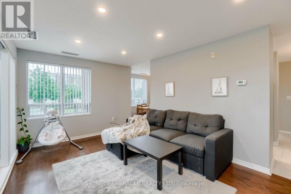 Living room with a dark grey sofa, coffee table, large windows, and wood floors. - 105 - 55 Via Rosedale Road, Brampton