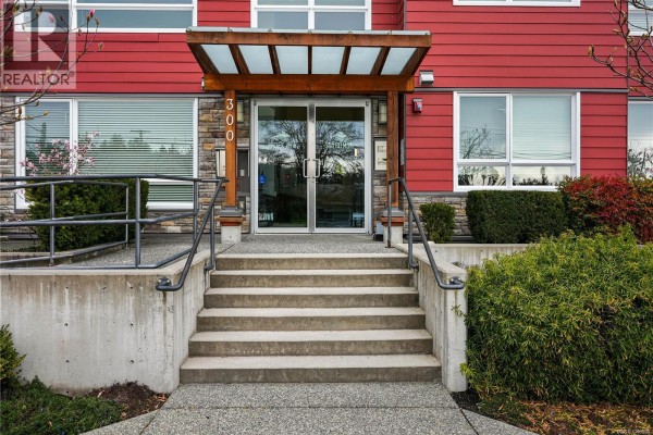 Bright living room with a sofa, side table, and large windows overlooking the street. - 105 300 Belmont Rd, Colwood