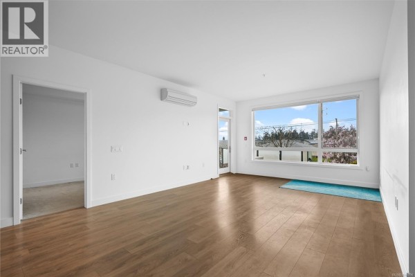 Empty living space with hardwood flooring and large window, featuring a view of the outdoors. - 105 300 Belmont Rd, Colwood