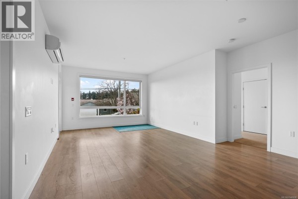 Another angle of the empty living room with a window and light-colored walls. - 105 300 Belmont Rd, Colwood