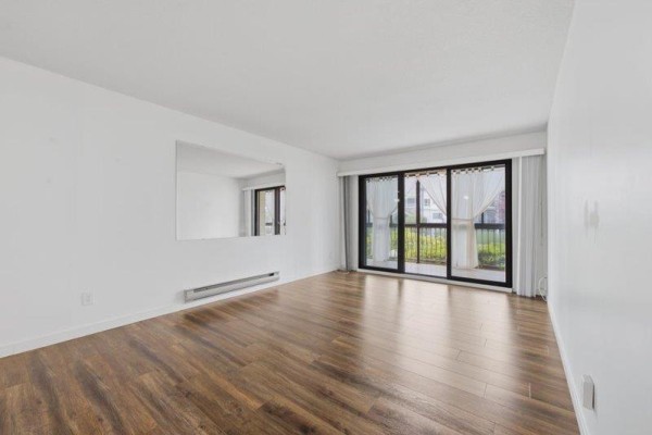 Empty living room with hardwood floors, large mirror, and sliding glass doors leading to a balcony. - 105 32033 Old Yale Road, Abbotsford