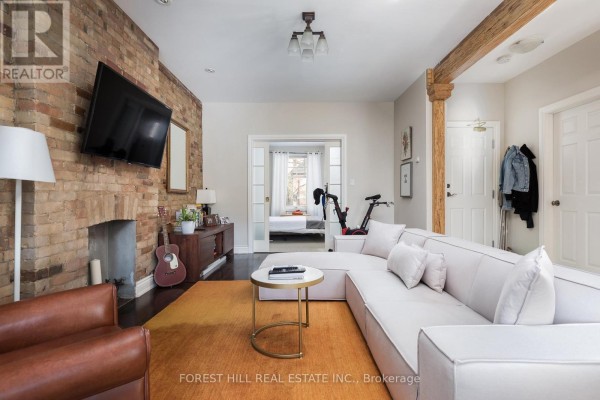 Living room with sectional sofa, brick fireplace, wooden beams, and an opening to a bedroom. - 105 Balmoral Avenue, Toronto