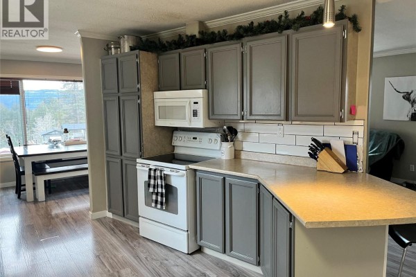 Kitchen with white stove, microwave, grey cabinets, and a dining area visible through the doorway. - 105 Jasper Drive, Logan Lake