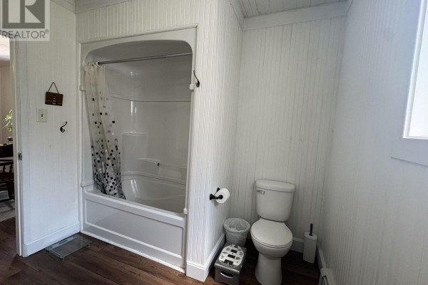 Small bathroom with white beadboard walls, toilet, bathtub with shower curtain, and dark wood-look flooring. - 105 Wilkes Cove Road, Middle Lahave