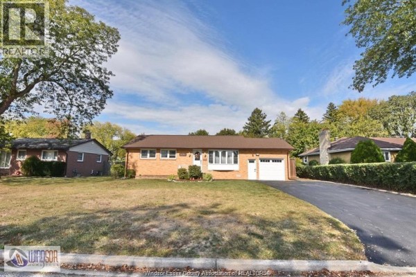 Exterior of a single-story brick house with a large lawn and mature trees. - 1050 Scofield, Windsor