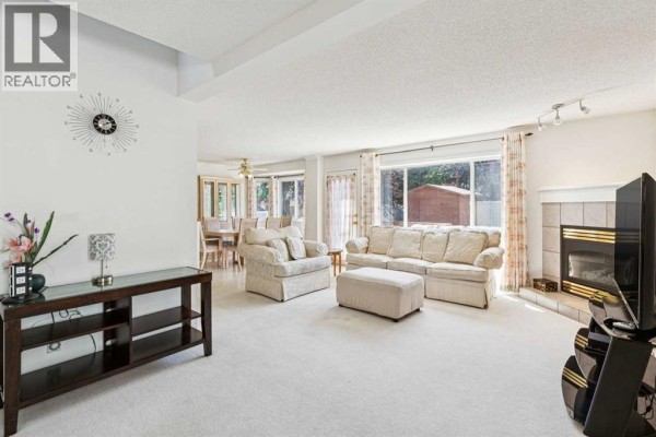 Living room with a fireplace, large windows overlooking a backyard, and a dining area visible in the background. - 10521 Hidden Valley Drive Nw, Calgary
