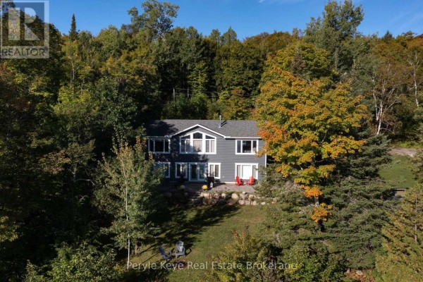 A two-story grey house with large windows is surrounded by trees with autumn foliage. - 1054 Inawendawin Road, Lake Of Bays