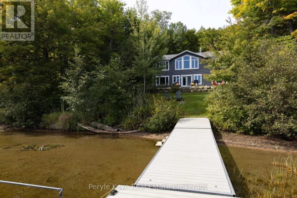 A grey dock extends from the shore towards a grey house nestled amongst trees. - 1054 Inawendawin Road, Lake Of Bays