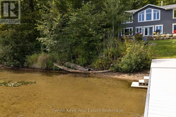 Clear lake water laps gently onto a sandy shore in front of a grey house. - 1054 Inawendawin Road, Lake Of Bays