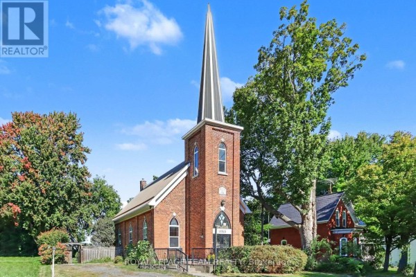 Front exterior of a red brick church with a prominent steeple and green trees. - 10565 Main Street, North Dundas