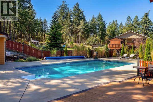 View of the backyard with a swimming pool, wooden deck, and surrounding trees. - 10590 Long Road, Lake Country